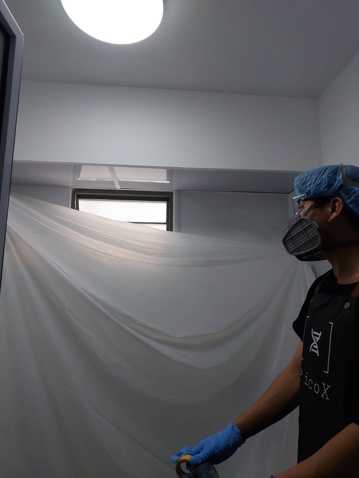 Person wearing protective gear taping a plastic sheet in a room with bright ceiling light.